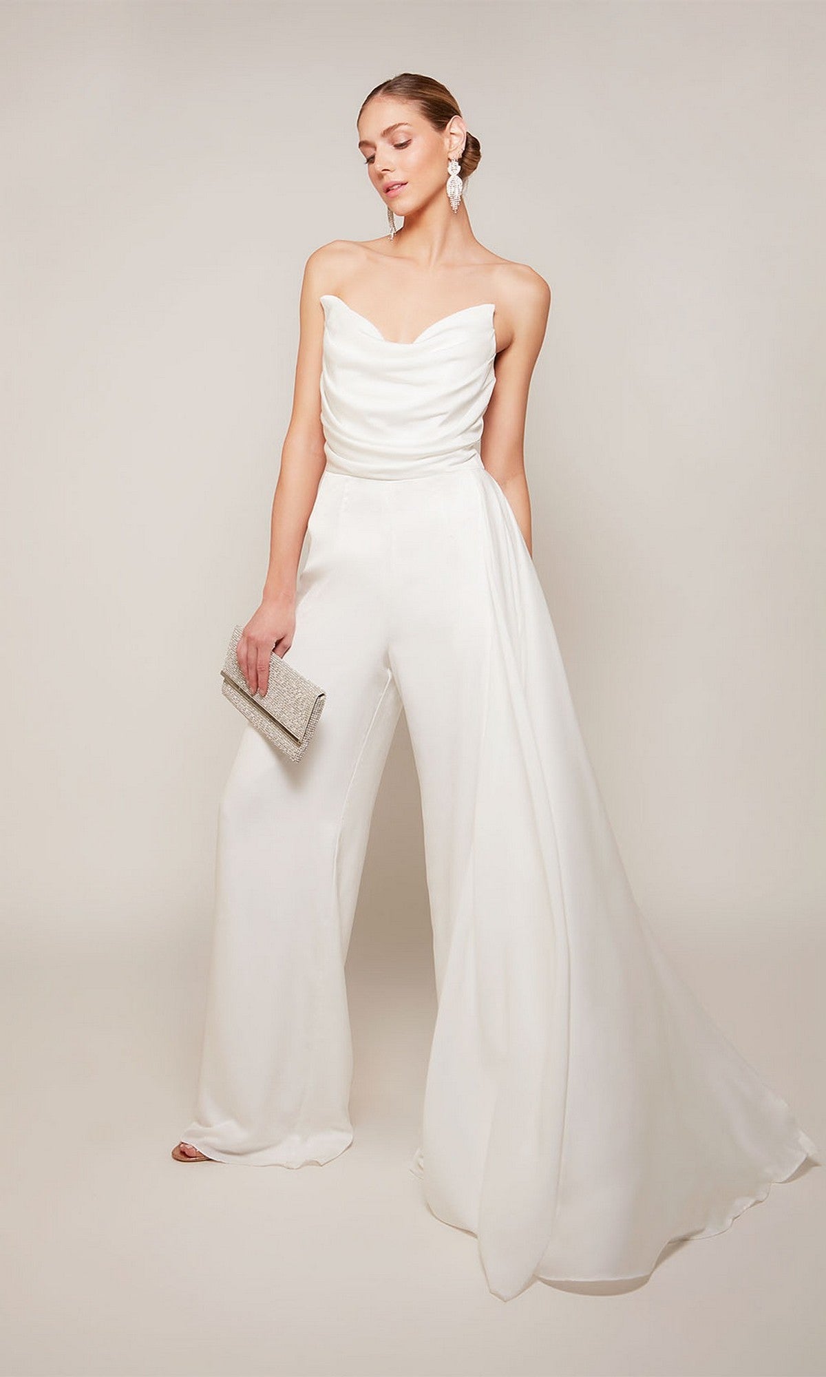 A woman wears the Alyce 70050 long formal jumpsuit featuring a strapless satin design with a draped bodice and wide legs, accessorized with a silver clutch and statement earrings.
